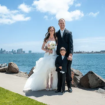 Bride and groom posing with young boy in formal wear by the waterfront on a sunny day.