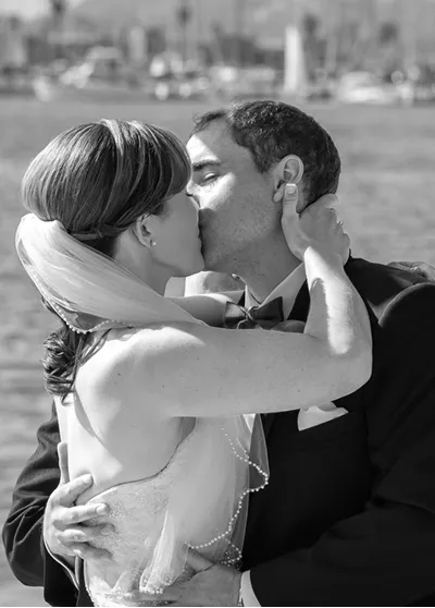 Bride and groom sharing a romantic kiss in front of a waterfront marina, with sailboats visible in the background.