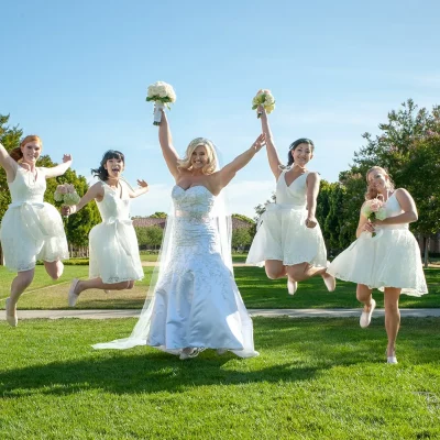Bride in white gown and bridesmaids in short white dresses jumping joyfully with bouquets outdoors
