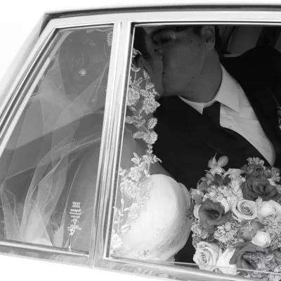 Bride and groom sharing a kiss inside a car, bride holding a bouquet of roses and wearing lace veil