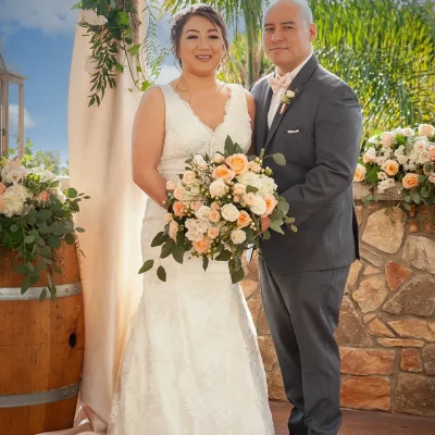 Bride and groom posing under a floral wedding arch with peach and white roses, bride holding bouquet