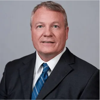 Professional man with short light hair wearing a dark suit, white shirt, and blue striped tie against a plain background