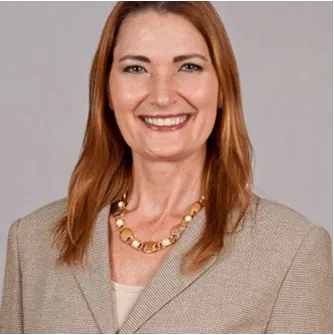 Confident professional woman with light brown hair wearing a beige business suit and gold necklace, smiling against a plain background