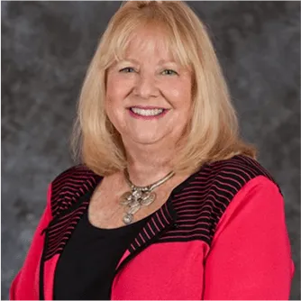 Smiling professional woman with blonde hair wearing a red blazer and necklace in front of a studio background