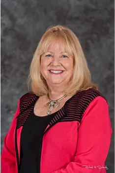 Smiling professional woman with blonde hair wearing a red jacket and necklace in front of a studio background