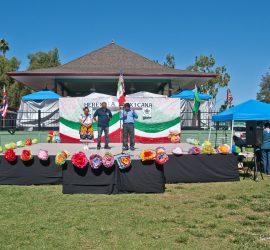 Two people stand on an outdoor stage decorated with colorful flowers and flags at a cultural event.