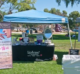A booth selling LipSense liquid lip color is set up in a park with promotional banners and display products on the table.