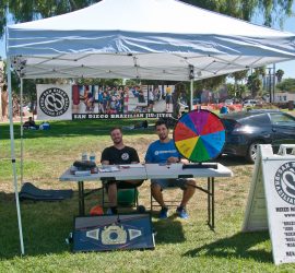 Two men sit at a San Diego Brazilian Jiu-Jitsu booth with a prize wheel, info board, and martial arts display at an outdoor event.
