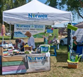 Norwex booth at an outdoor event displaying cleaning products, informational banners, and staffed by people under a white tent.