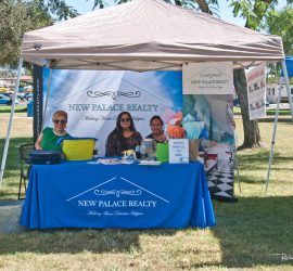 Three people sit at a New Palace Realty booth under a canopy in a park, with marketing materials on the table.