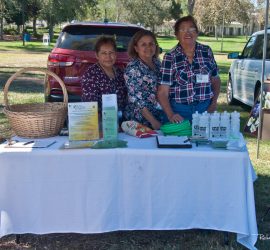 Three women stand behind a table with pamphlets, water bottles, and baskets at an outdoor park event.