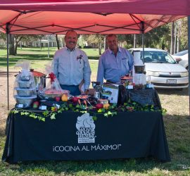 Two men stand behind a table with kitchen items and food samples under a red canopy at an outdoor event.