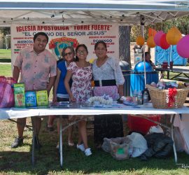 Four people stand behind a table with food and drinks at a church booth in a park, with balloons and a banner behind them.