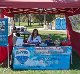 A woman and child sit at a real estate booth in a park, with brochures and a sign for RE/MAX United on the table.