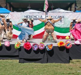 Mexican folkloric dancers perform on an outdoor stage decorated with large flowers and a sign reading “Herencia Mexicana.”.