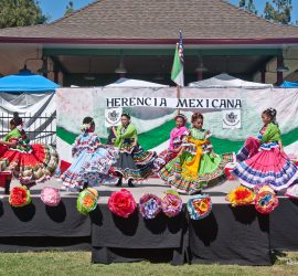 Five girls in colorful traditional Mexican dresses dance on an outdoor stage decorated with paper flowers and a "Herencia Mexicana" banner.