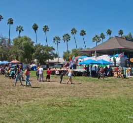 Outdoor community event with people, tents, and balloons on a sunny day near a historic house with American flags.