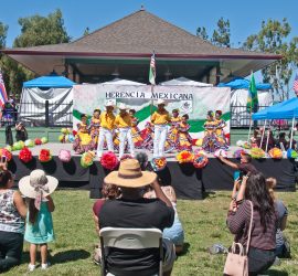 Dancers in traditional Mexican attire perform on an outdoor stage as audience members watch and take photos.