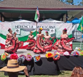 A group of girls in traditional Mexican dresses perform a folkloric dance on an outdoor stage decorated with flowers.