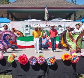 Five people in traditional Mexican attire perform a folkloric dance on an outdoor stage decorated with paper flowers.