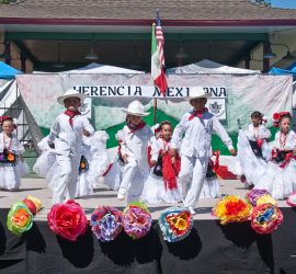 Children dressed in traditional Mexican attire perform a folkloric dance on an outdoor stage decorated with paper flowers.
