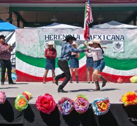 Group of people in cowboy attire dance on stage with a "Herencia Mexicana" banner and paper flowers in front.