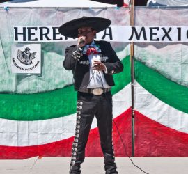 A man in traditional charro attire sings on stage at a Mexican heritage event, with colorful paper flowers in front.