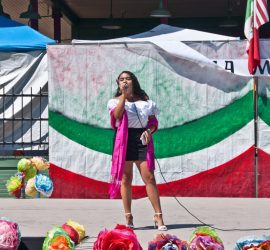 Woman in traditional attire sings on an outdoor stage decorated with colorful flowers and national flags.