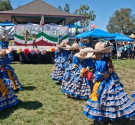 Young girls in blue traditional dresses perform a folkloric dance outdoors during a Mexican cultural event.