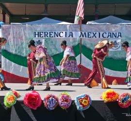 Six dancers in traditional Mexican attire perform on stage in front of a "Herencia Mexicana" banner and decorations.