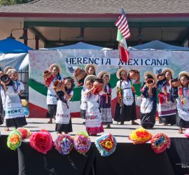 Children in traditional Mexican attire perform a folk dance on stage with "Herencia Mexicana" banner in the background.