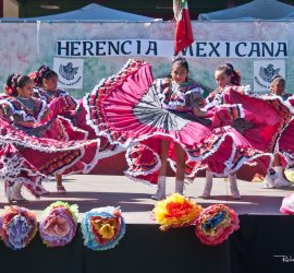 Young girls in colorful traditional dresses perform a Mexican folk dance on stage at a cultural event.