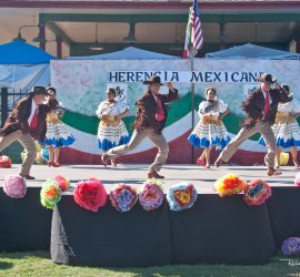 Group of dancers in traditional Mexican attire perform on an outdoor stage decorated with flowers and a "Herencia Mexicana" banner.