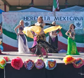 Six women perform a belly dance on an outdoor stage decorated with flowers and a sign reading "Herencia Mexicana.