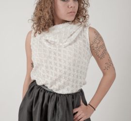 Person with curly hair wearing a white textured top and dark skirt, posing against a plain light background.