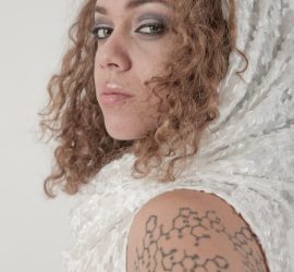 Woman with curly hair in a white sequin hood, showing a large black geometric tattoo on her upper arm.