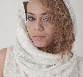 Woman with curly hair wearing a textured white hooded garment, looking at the camera against a plain background.