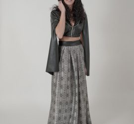Woman standing in a studio wearing a hooded black crop top and a long patterned skirt, with a neutral background.