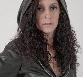 Woman with long curly hair wearing a black hooded leather jacket, looking at the camera against a plain background.