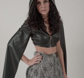 Woman wearing a black hooded crop top and patterned pants stands against a plain background, looking at the camera.