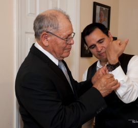 An older man in a suit helps a younger man adjust his cuff in a room with framed photos on the wall.