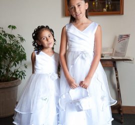 Two young girls in white formal dresses stand indoors, posing for a photo in front of a wooden table and plant.