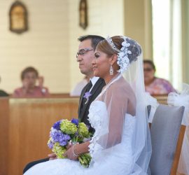 A bride and groom sit side by side in a church, the bride holding a bouquet of flowers, during a wedding ceremony.