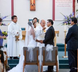 Bride and groom holding hands at the altar during a church wedding ceremony with priest and wedding party present.