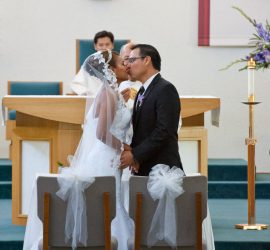 Bride and groom kiss at the altar during a wedding ceremony in a church, with a priest in the background.