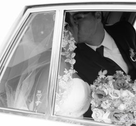 A bride and groom share a kiss inside a car, with the bride holding a bouquet and wearing a lace veil.