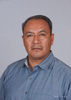 A man with short dark hair wearing a blue patterned collared shirt poses against a plain light background.