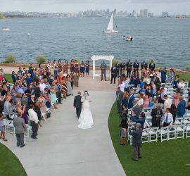 A bride walks down the aisle with a man at an outdoor wedding by the water, guests seated and standing on both sides.