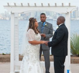A bride and groom stand facing each other, holding hands, during an outdoor waterfront wedding ceremony with an officiant.