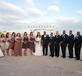 A wedding party poses outdoors near water, with bridesmaids in brown dresses and groomsmen in black suits.